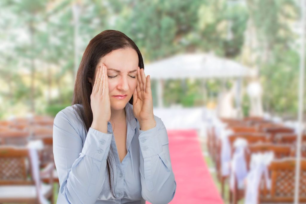 Female venue owner in the foreground with eyes closed and hands at her temples, showing frustration. In the background, the wedding ceremony area with chairs and an altar is visible.